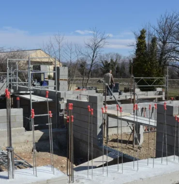 Budowa domu jednorodzinnego na etapie fundamentów: betonowy fundament z wystającymi prętami zbrojenia i ściany z bloczków betonowych, w tle rusztowania i bezlistne drzewa