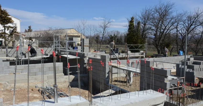 Budowa domu jednorodzinnego na etapie fundamentów: betonowy fundament z wystającymi prętami zbrojenia i ściany z bloczków betonowych, w tle rusztowania i bezlistne drzewa